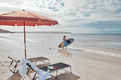 Two beach chairs under a red umbrella on a sandy beach, with a surfer walking along the shoreline under a cloudy sky, capturing the tranquil experience of luxury escapes at CABN Kangaroo Island